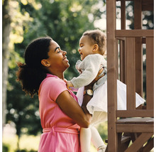 Femme tenant un jeune enfant devant une tour de jeux en bois dans le jardin.