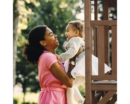Femme tenant un jeune enfant devant une tour de jeux en bois dans le jardin.