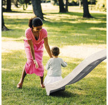 Femme et enfant jouant avec un toboggan de jardin en plastique gris clair sur une pelouse verte dans un environnement lumineux et spacieux.