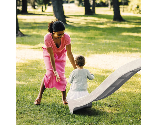 Femme et enfant jouant avec un toboggan de jardin en plastique gris clair sur une pelouse verte dans un environnement lumineux et spacieux.