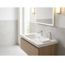 Helles Badezimmer mit weißem Waschtisch auf Holzunterschrank, verchromter Armatur, beleuchtetem Spiegel und Wandleuchten.