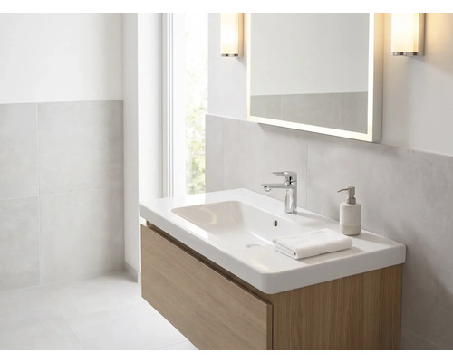 Helles Badezimmer mit weißem Waschtisch auf Holzunterschrank, verchromter Armatur, beleuchtetem Spiegel und Wandleuchten.