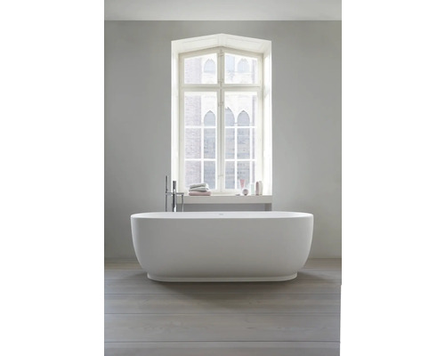 Baignoire îlot blanche dans une salle de bain moderne et lumineuse devant une fenêtre, robinetterie au sol, parquet.