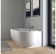 Baignoire ovale moderne blanche avec robinetterie murale chromée dans une salle de bains lumineuse avec murs aspect béton et parquet.