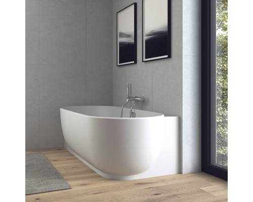 Baignoire ovale moderne blanche avec robinetterie murale chromée dans une salle de bains lumineuse avec murs aspect béton et parquet.