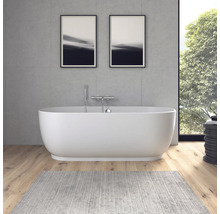 Baignoire îlot ovale moderne devant un mur aspect béton, parquet et tapis gris dans une salle de bains lumineuse.