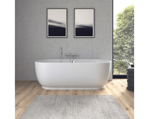 Baignoire îlot ovale moderne devant un mur aspect béton, parquet et tapis gris dans une salle de bains lumineuse.