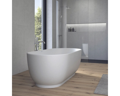 Baignoire îlot ovale dans une salle de bains moderne avec des murs aspect béton, un sol en bois et une douche à l'arrière-plan.