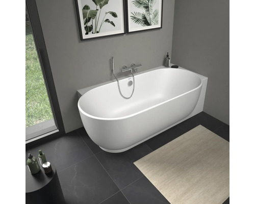 Baignoire en îlot blanche moderne de forme ovale dans une salle de bains lumineuse avec robinetterie murale chromée et carrelage foncé.