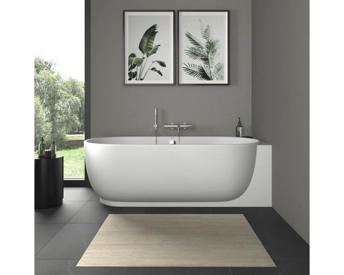 Salle de bains moderne avec baignoire en îlot ovale blanche, robinetterie murale, tableaux botaniques et tapis de bain clair.