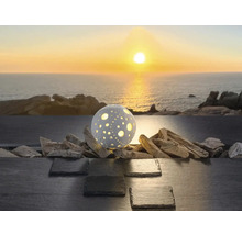 Kugelförmige dekorative Außenleuchte mit Lochmuster auf Treibholz und Schiefer vor dem Meer bei Sonnenuntergang.