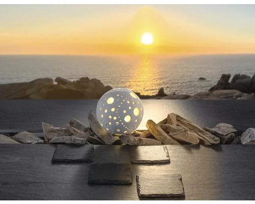 Kugelförmige dekorative Außenleuchte mit Lochmuster auf Treibholz und Schiefer vor dem Meer bei Sonnenuntergang.
