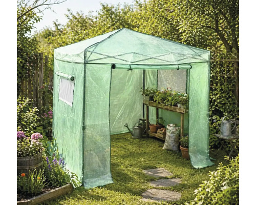 Serre de jardin en film plastique avec étagère en bois, plantes en pots, arrosoir et sac de terreau sur l herbe.