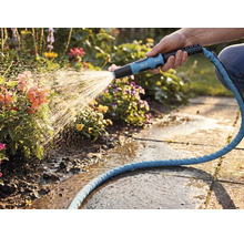 Eine Hand bewässert mit einer for_q Garten-Spritzdüse und einem blauen Textilschlauch Blumen in einem hellen Garten mit Steinplatten.