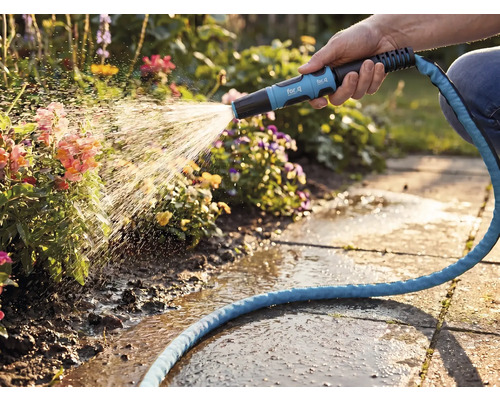 Eine Hand bewässert mit einer for_q Garten-Spritzdüse und einem blauen Textilschlauch Blumen in einem hellen Garten mit Steinplatten.