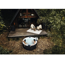 Des personnes se détendent dans un spa gonflable rond à l'extérieur devant une maison en bois en forme de A avec une terrasse et des guirlandes.