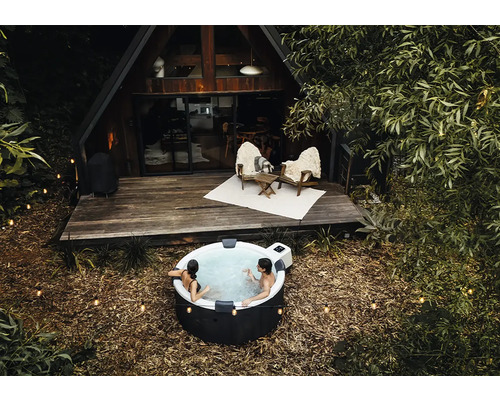 Des personnes se détendent dans un spa gonflable rond à l'extérieur devant une maison en bois en forme de A avec une terrasse et des guirlandes.
