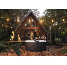 Jacuzzi extérieur rond dans un jardin avec une maison moderne en forme de A, des guirlandes lumineuses et des rennes décoratifs le soir.