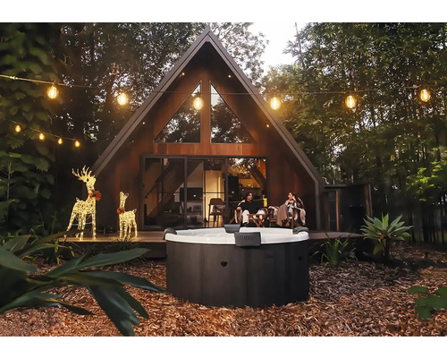 Jacuzzi extérieur rond dans un jardin avec une maison moderne en forme de A, des guirlandes lumineuses et des rennes décoratifs le soir.