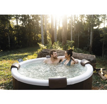 Un couple se détend dans un jacuzzi extérieur rond dans un jardin avec des arbres. Le jacuzzi dispose de repose-tête et d'eau bouillonnante.