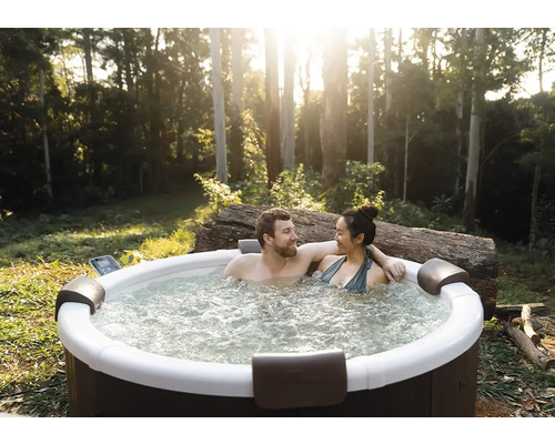 Un couple se détend dans un jacuzzi extérieur rond dans un jardin avec des arbres. Le jacuzzi dispose de repose-tête et d'eau bouillonnante.