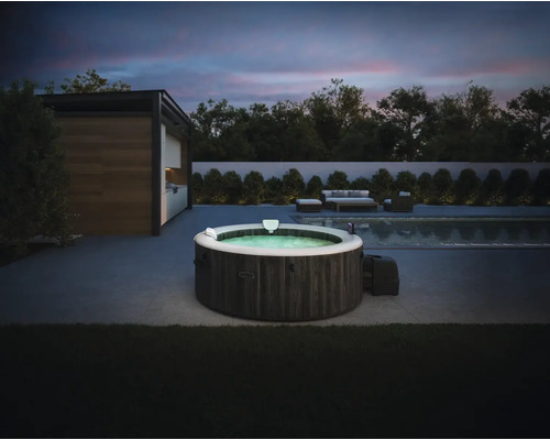 Jacuzzi gonflable Intex aspect bois foncé sur une terrasse moderne à côté d une piscine au crépuscule avec mobilier de jardin en arrière-plan.