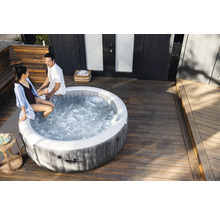 Un spa gonflable rond avec aspect bois sur une terrasse en bois clair avec deux personnes assises sur le rebord.
