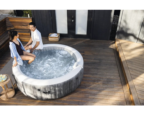 Un spa gonflable rond avec aspect bois sur une terrasse en bois clair avec deux personnes assises sur le rebord.