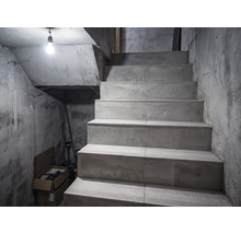 Escalier en béton dans une cave aux murs bruts, éclairé par une seule ampoule.