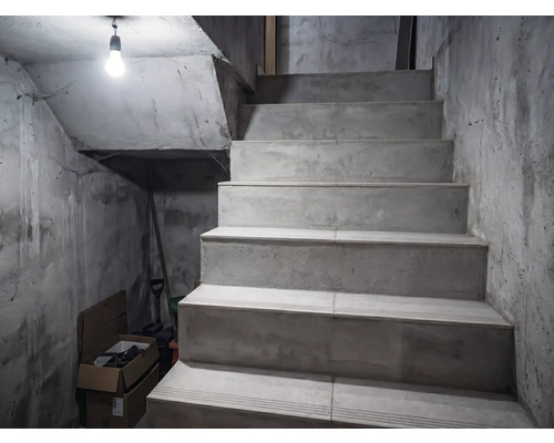 Escalier en béton dans une cave aux murs bruts, éclairé par une seule ampoule.