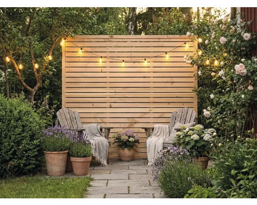 Paravent en bois à lattes horizontales, deux chaises en bois avec couvertures, pots en terre cuite avec lavande et hortensias et une guirlande lumineuse.