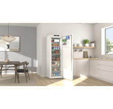 Congélateur blanc avec porte ouverte dans une cuisine moderne et lumineuse avec coin repas et aliments bien rangés.