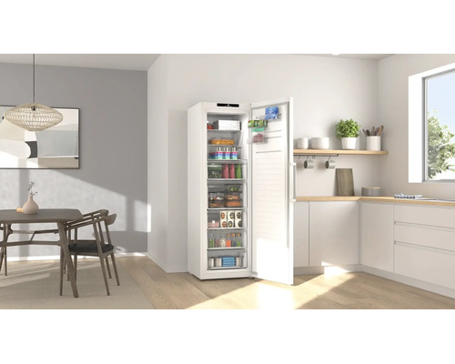 Congélateur blanc avec porte ouverte dans une cuisine moderne et lumineuse avec coin repas et aliments bien rangés.
