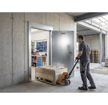 Homme tirant un transpalette avec caisse en bois par une porte de sécurité métallique Abus ouverte dans un bâtiment en béton avec atelier.
