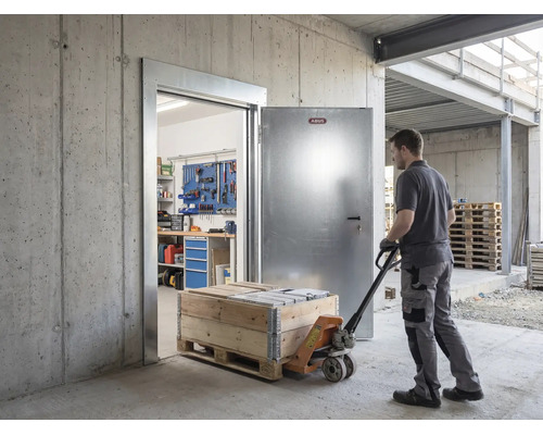 Homme tirant un transpalette avec caisse en bois par une porte de sécurité métallique Abus ouverte dans un bâtiment en béton avec atelier.