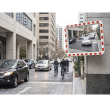 Rechteckiger Verkehrsspiegel mit rot-weißem Rahmen an einer Wand, der eine städtische Straße mit Autos reflektiert.
