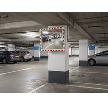 Rechteckiger Verkehrsspiegel mit rot-weißem Rahmen, montiert an einer Betonsäule in einer hellen, organisierten Tiefgarage mit geparkten Autos.