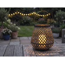 Lanterne tressée avec bougie LED sur une table en bois dans un jardin au crépuscule avec guirlande lumineuse en arrière-plan.