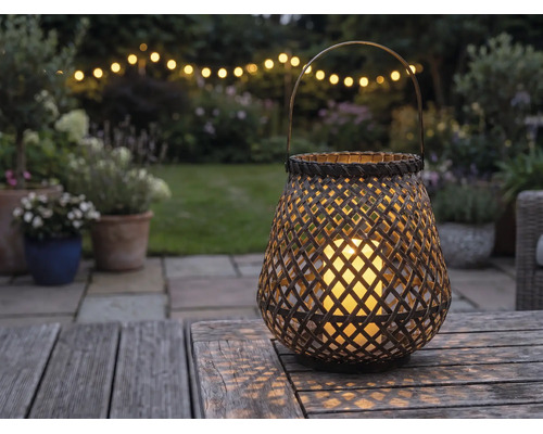 Lanterne tressée avec bougie LED sur une table en bois dans un jardin au crépuscule avec guirlande lumineuse en arrière-plan.