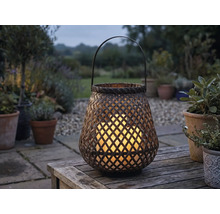 Lanterne de jardin tressée en bambou avec bougies LED allumées sur une table en bois dans un jardin au crépuscule.