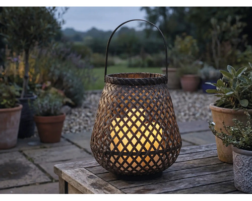 Lanterne de jardin tressée en bambou avec bougies LED allumées sur une table en bois dans un jardin au crépuscule.