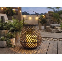 Lanterne de jardin tressée avec anse et bougie allumée sur une terrasse en bois parmi des plantes vertes.