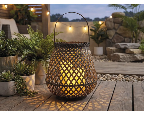 Lanterne de jardin tressée avec anse et bougie allumée sur une terrasse en bois parmi des plantes vertes.