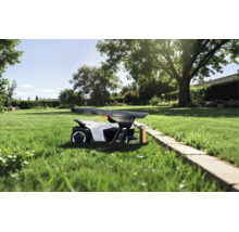 Mähroboter unter einer Garagenabdeckung auf einer Rasenfläche in einem sonnigen Garten mit Bäumen und Steineinfassung.