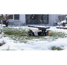 Mähroboter in einer Roboter-Garage auf einer verschneiten Rasenfläche während Schneefall vor einem Haus.