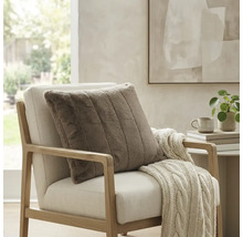 Coussin décoratif carré côtelé en fausse fourrure sur un fauteuil clair avec un plaid en tricot dans une pièce lumineuse et moderne.