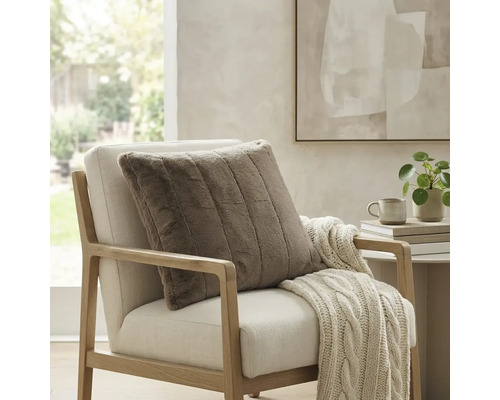 Coussin décoratif carré côtelé en fausse fourrure sur un fauteuil clair avec un plaid en tricot dans une pièce lumineuse et moderne.