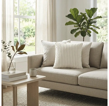 Helles Wohnzimmer mit beigem Stoffsofa, weißem Cord-Zierkissen und Holz-Beistelltisch. Große Zimmerpflanze und Fenster im Hintergrund.