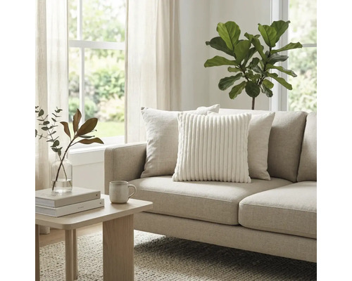 Helles Wohnzimmer mit beigem Stoffsofa, weißem Cord-Zierkissen und Holz-Beistelltisch. Große Zimmerpflanze und Fenster im Hintergrund.