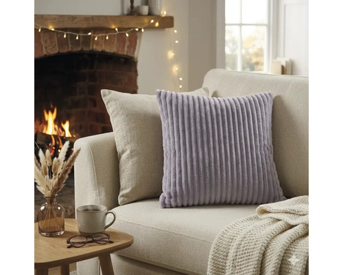 Coussin décoratif lavande à texture côtelée sur un canapé clair dans un salon chaleureux avec cheminée allumée et table d'appoint.